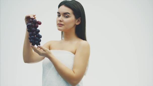 Menina bonita jovem de pé sobre um fundo branco. Durante isso, ele segura as uvas em suas mãos e olha para ele olhando para a câmera . — Vídeo de Stock