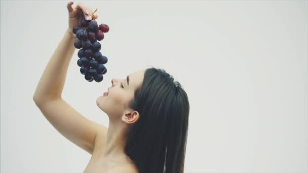 Uma menina bonita e magra come frutas saudáveis. Retrato de uma jovem bonita segurando um buquê de uva madura e uma verdade. Durante este tempo é sobre um fundo branco . — Vídeo de Stock