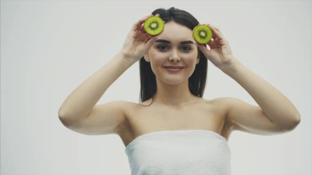 Mladá hezká dívka stojící na bílém pozadí. Během tohoto na úrovni očí, kiwi částice jsou uchovávány v rukou zavřenýma očima zobrazující jazyk. — Stock video