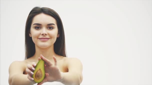 Avocado voor een gezond lichaam. Mooi jong meisje bevat een halve avocado in haar handen en wordt op de camera. Gedurende deze tijd staat het op een witte achtergrond. Mooie lange zwarte haren. — Stockvideo
