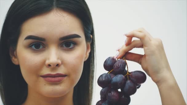 Jeune jolie fille debout sur un fond blanc. Pendant ce temps, il regarde la caméra et lève la main avec des raisins, qui est sur un fond flou. Au bout d'un moment, il tombe. A beau — Video