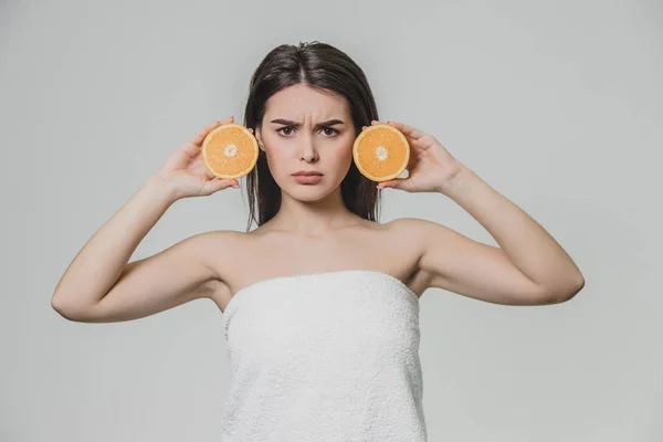 Chica joven y feliz posa con fruta. Durante esto ella mira a la cámara. Sostiene dos piezas de naranja en sus manos y es muy grave . — Foto de Stock