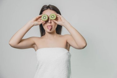 Genç güzel kız beyaz bir arka plan üzerinde ayakta. Bu sırada, Dil Gösterilen kapalı gözler elinde kivi parçacıklar göz düzeyinde tutulur.