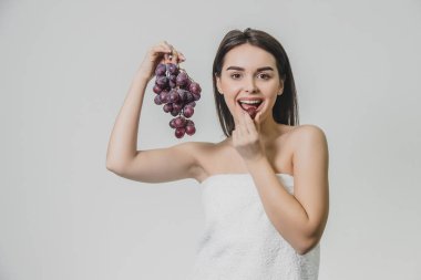 Güzel genç esmer kız mutlu bir gülümseme ile. Taze üzüm demet elinde tutarak, beyaz bir arka plan üzerinde izole. Bu süre içinde o kameraya bakar ve üzüm yiyor.
