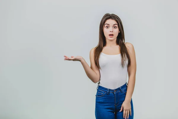 Young pretty Caucasian girl standing on a white background. During this time, wearing a white T-shirt and blue jeans. Shows his hand to the side and looks at the camera. Has beautiful long black hair. — Stock Photo, Image