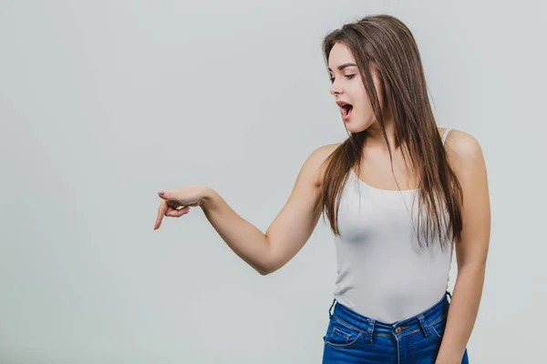 A young pretty Caucasian girl stood on a white background. During this time wear a white T-shirt and blue jeans. Shows the index finger on the floor and opens the mouth to the finger. — Stock Photo, Image