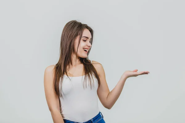 A young pretty Caucasian girl stood on a white background. During this time wear a white T-shirt and blue jeans. Turning his hand to the side looks at her. — Stock Photo, Image