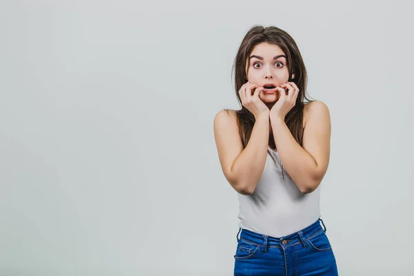 Giovane bella ragazza in piedi su uno sfondo bianco. Durante questo, lei è stupita di mettere le mani sul suo viso e fissa con timore nella direzione. Ha bei capelli lunghi neri . — Foto Stock