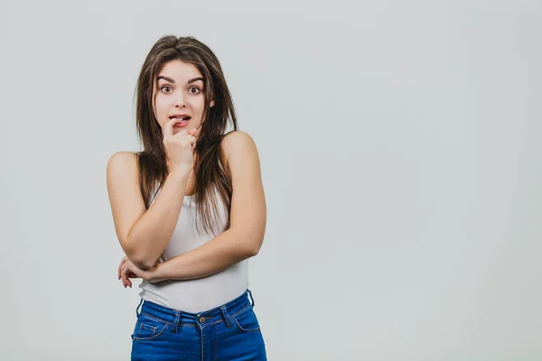 Menina bonita jovem de pé sobre um fundo branco. Durante isso, ela coloca um dedo em sua boca e olha para a câmera de surpresa. Tem cabelo longo preto bonito . — Fotografia de Stock