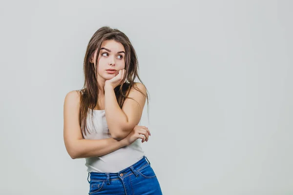 Une jolie jeune fille debout sur un fond blanc. La fille est vêtue d'une chemise blanche. A de beaux cheveux longs noirs. Après réflexion, elle mit sa main sous le menton. Regardez sur le côté . — Photo