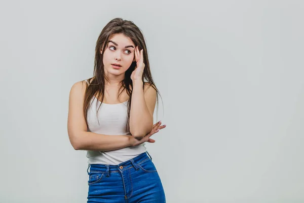 Primer plano de una chica joven y bonita de pie sobre un fondo blanco. La chica está vestida con una camisa blanca y un jersey azul. Tiene hermoso pelo largo negro. Sorprendido al poner una mano sobre su cabeza la otra —  Fotos de Stock