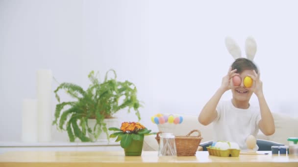 Lindo niño con orejas de conejo sosteniendo huevos de Pascua rosados y amarillos imitándolo como ojos. Pequeño, divertido, travieso chico está mostrando su toungue . — Vídeos de Stock