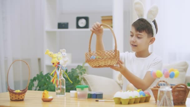 Милый маленький мальчик с кроличьими ушами держит деревянную корзину, полную пасхальных красочных яиц, и показывает пальцем, как сильно он любит пасхальное зачатие . — стоковое видео