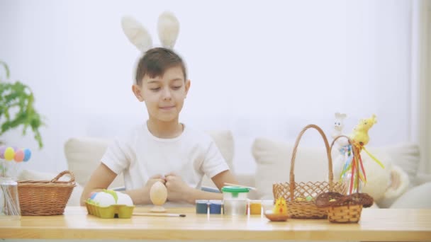 Söt liten pojke med bunny öron är vid bordet fullt av påskpynt och höjer fingrarna tummen upp, leende. Söt påsk pojke tar en naken borste och börjat måla en påsk — Stockvideo