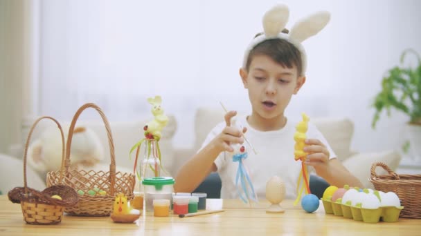 Der kleine süße Junge sitzt am Tisch voller Osterdekorationen und spielt mit Osterhasen in seinen Händen. Hasentheater. — Stockvideo