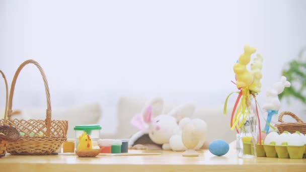 Little boy is hiding under the wooden table, full of Easter decorations: basket, yellow chicken, colorful eggs pains and paint-brush. Boy is playing with a cute, soft white bunny with pink ears, on — Stock Video