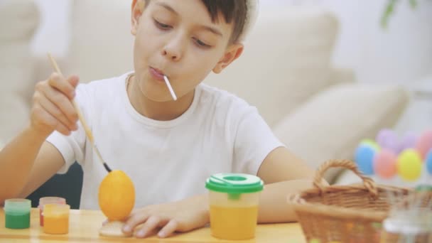 Заинтересованный и сосредоточенный мальчик заканчивает раскрашивать пасхальное яйцо желтым цветом, сидя за столом и пробуя сладкий леденец . — стоковое видео