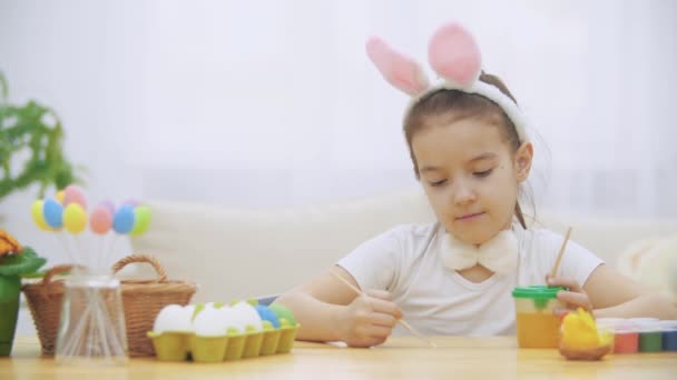 Little cute girl is having fun holding a paint-brush in her left hand. Girl with a beauty spots at her face watching at nude paint-brushes, sitting at the wooden table with Easter decorations. Girl is — Stock Video