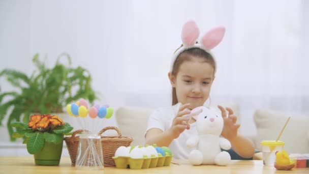 Brisk, cute girl is having fun painting with Easter bunny, which has the same ears, as she has. Girl is setting back white bunny. Girl with a beauty spot at her face and is smiling widely, sitting at — Stock Video