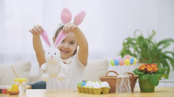 Little cute girl is having fun painting with Easter bunny, which has the same ears, as she has. Smiling girl is expanding bunnys ears and then is hugging it. Girl with a beauty spot at her face and — Stock Video