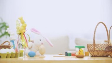 Küçük çocuk ahşap masanın altından Paskalya süsleri tam gizleme: sepet, sarı tavuk, renkli yumurta ağrıları ve boya fırçası. Çocuk şirin, yumuşak beyaz tavşan pembe kulaklı ile oynuyor