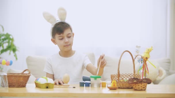Ragazzo creativo sta colorando un uovo di Pasqua con un aiuto di pennello., indossando orecchie di coniglio, poi alza le dita del pollice . — Video Stock