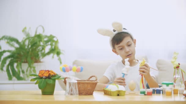 Jovem menino adorável está sentado à mesa cheia de decorações de Páscoa e está brincando com coelhos de Páscoa em suas mãos. Discussão de coelhinhos. Quem pintaria um teatro de ovo Bunny . — Vídeo de Stock