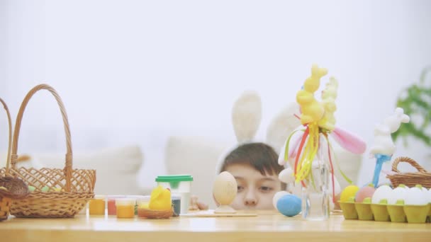 Playful boy is hiding under the wooden table, full of Easter decorations: basket, yellow chicken, colorful eggs pains and paint-brush. Boy is playing with a cute, soft white bunny with pink ears, on — Stock Video
