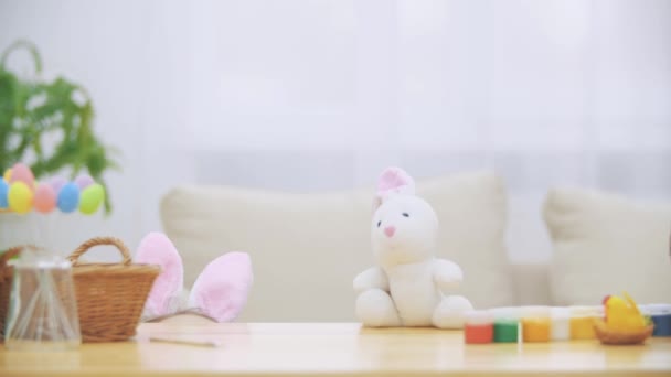Petite fille mignonne avec des oreilles de lapin se cache sous la table pleine de décorations de Pâques. Petit lapin blanc mignon attaque fille gentiment. Rire est la chambre . — Video