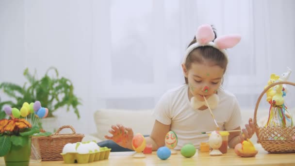 Menina bonito está se divertindo pintura. Menina adorável é colorir um ovo de Páscoa e suas mãos com uma ajuda de pincel de tinta em sua boca. Menina com manchas de beleza em seu rosto e está olhando suavemente com gatos — Vídeo de Stock