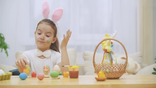 Pequena menina bonito e adorável está sorrindo sinceramente. Ela pega um ovo de Páscoa e mostra o resultado de seu trabalho, usando suas mãos sujas de tintas. A rapariga está a acenar . — Vídeo de Stock