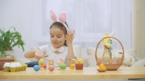 Litlte menina brincalhão, vestindo orelhas de coelho em sua cabeça está pintando suas mãos. De repente, alguém reparou nela. Ela ficou assustada e escondeu as mãos. . — Vídeo de Stock