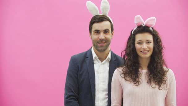 Young beautiful couple standing on a pink background. With hackneyed ears on the head. During this man and his wife are looking into the camera. — Stock Video