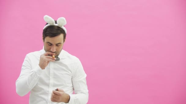 Young handsome boy standing on a pink background. A man with rabble ears on his head. A man holding a carrot puts it in his mouth and wants to light an ashtray. Throws carrots and taking a cigar lit — Stock Video