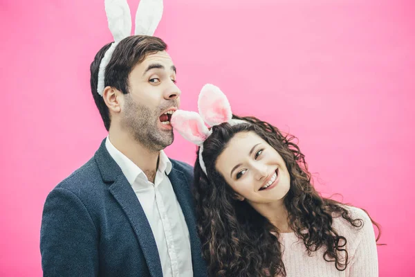 Young happy couple standing on pink background. With a bunny ears on the head. The girl put her head on the mans shoulder, looking at the camera smiling. He opened his mouth to take her ear in his