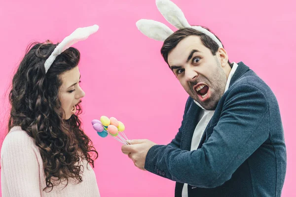 Young happy family on a pink background. At the same time on the head there is a rabbits ears. During this man holding decorative eggs in his hands, he wants to put them in the mouth of his wife. It — Stock Photo, Image