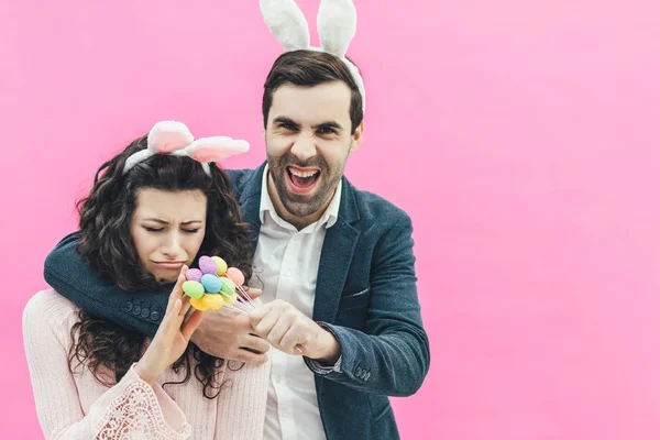 Junge glückliche Familie auf rosa Hintergrund. Gleichzeitig befindet sich auf dem Kopf ein Hasenohren. , während dieser Mann dekorative Eier in den Händen hält und seine Frau lächelnd umarmt. Ostern. — Stockfoto