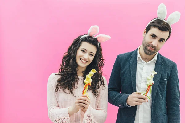 Young pretty sweet couple standing on a pink background. At the same time, there are hooves on the head. A husband and wife look at the camera carefully while holding sweet birds in their hands