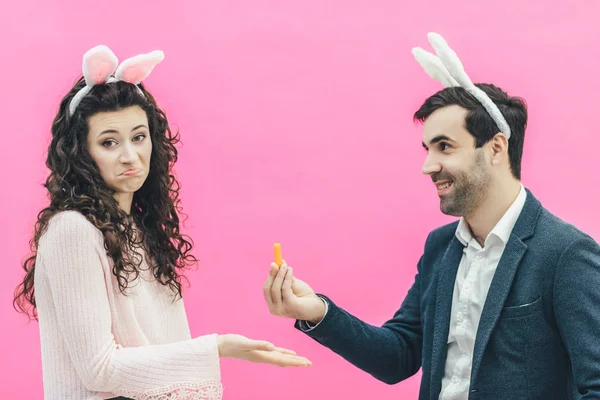 Молода щаслива пара на рожевому тлі. На голові кролячі вуха. Молодий чоловік, який тримає маленьку моркву в руках, показує свою дружину. Дівчина здивована, що поклала руку в горизонтальне положення — стокове фото