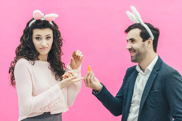Young happy couple on the pink background. On the head is a rabbit ears. A young man holding a carrot of small size in his hands shows dorkuzhina. The girl in surprise shows her index finger on — Stock Photo, Image