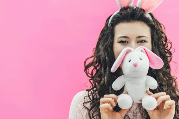 Junges hübsches Mädchen, das auf rosa Hintergrund steht. auf dem Kopf der Ohren des Hasen. Dabei hält er eine weiche Hasenhand in seinen Händen und legt sein Gesicht in den Hintergrund. Ostern. — Stockfoto