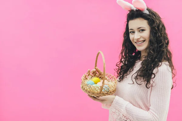 Zbliżenie: młode, piękne dziewczyny, stojącą na tle różowej. Na głowie są uszy królika. W tym samym czasie uśmiecha się i posiada kolorowy kosz Wielkanoc jaja w jego ręce. Patrząc na — Zdjęcie stockowe