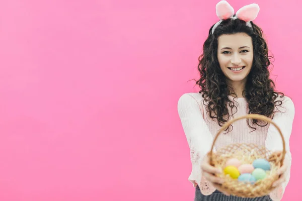 Młode ładne dziewczynę stojącą na tle różowej. Na głowie są uszy królika. W tym samym czasie uśmiecha się i posiada kolorowy kosz Wielkanoc jaja w jego ręce. Zajrzeć do aparatu. — Zdjęcie stockowe