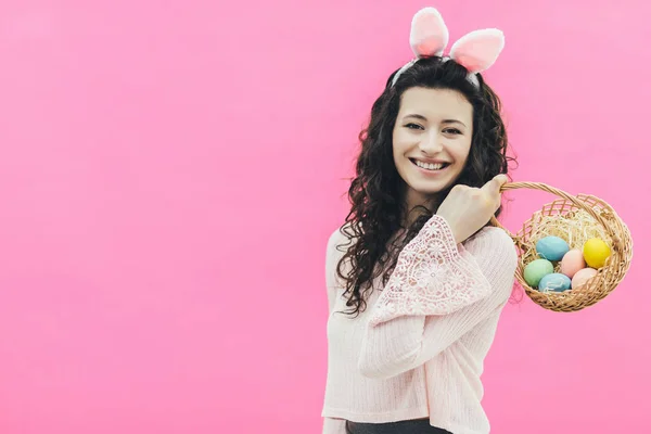 Młoda Ładna dziewczyna na różowym tle. Na głowie są różowy króliczek uszu. Szczery uśmiech, trzymając kosz z dekoracyjne jajka w jego ręce, oddanie jego ramię. Poradzić sobie Cpase. Wielkanoc. — Zdjęcie stockowe