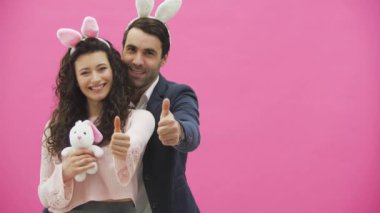 Genç pembe bir arka plan üzerinde duran çift. Bu gösterisi sırasında bir jest sınıf gülümseyen ve yumuşak oyuncak tavşan elinde tutan. Mutlu bir aile bir pembe kulakları ile Paskalya için hazırlanıyor