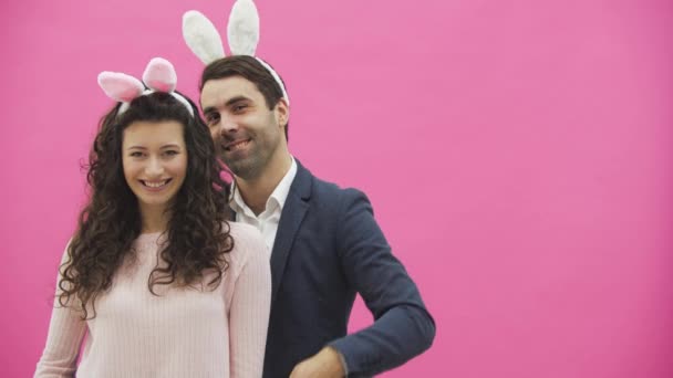 젊은 커플 핑크 배경에 서 있는. 이, 그들은 카메라에 모양과 전화의 제스처를 보여. 행복 한 가족은 그것의 머리에 분홍색 토끼의 귀와 부활절 준비. — 비디오
