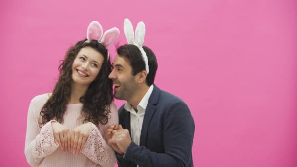 Schönes junges Paar auf rosa Hintergrund stehend. Während dieser Zeit stellen sie die Bewegungen von Hasen nach und legen ihre Hände wie ein Kaninchen. zusammen mit rosa bellenden Ohren auf dem Kopf. glückliche Familie — Stockvideo