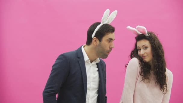 Hermosa pareja joven de pie sobre fondo rosa. Al mismo tiempo, la niña aplaude a su marido, él a su vez le muerde la mano. Junto con las orejas de color rosa ladrando en la cabeza. Familia feliz se está preparando para — Vídeos de Stock