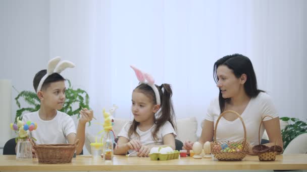 Mutter ist mit ihren Kindern da: Tochter und Sohn, die Hasenohren tragen, am Tisch sitzen und Pinsel in der Hand halten. Tochter versucht, eine orangefarbene Farbe zu öffnen. Zeitlupe. — Stockvideo