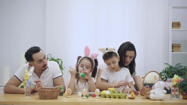 Les parents avec leurs adorables et mignons enfants, qui leur ressemblent vraiment, colorent les œufs de Pâques, assis à la table en bois, plein de décorations de Pâques. La famille est concentrée sur l'activité — Video